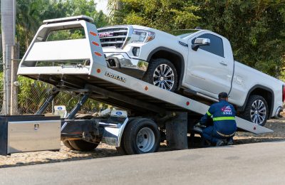 Tow truck operator loading white GMC pickup truck on street in daytime.